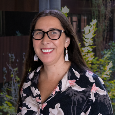 Headshot of Stephanie Carroll wearing glasses, white earrings, and a floral button-down shirt with a dark background.